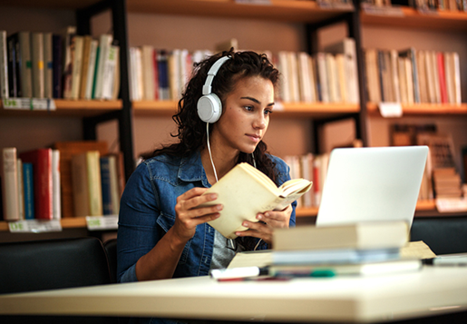 Girl in library holding book and looking at laptop while wearing headphones.